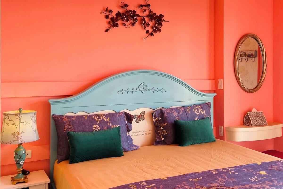 A bedroom with a bed, a nightstand, a lamp, a mirror, and a wall decoration.