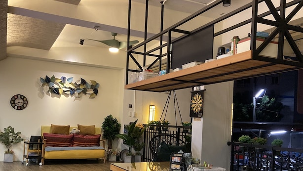 Een moderne woonkamer met een houten bank, een sofa en een eettafel met stoelen. Er is een plafondventilator, een klok en een dartbord aan de muur.