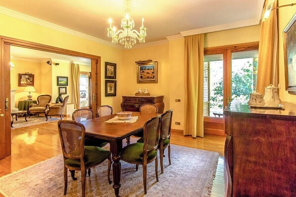 A dining room with a wooden table and chairs, a chandelier, framed pictures on the walls, and a view of the outdoors through large windows.