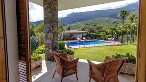 A patio with wicker chairs, a stone pillar, and a view of a swimming pool and house in a lush landscape. - Mountain view villa with private pool near Costa Brava (Gerona / Girona)