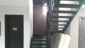 Un escalier avec une main courante et des marches en bois foncé mène à un palier où se trouve un luminaire. Une porte avec une enseigne est visible sur la gauche.