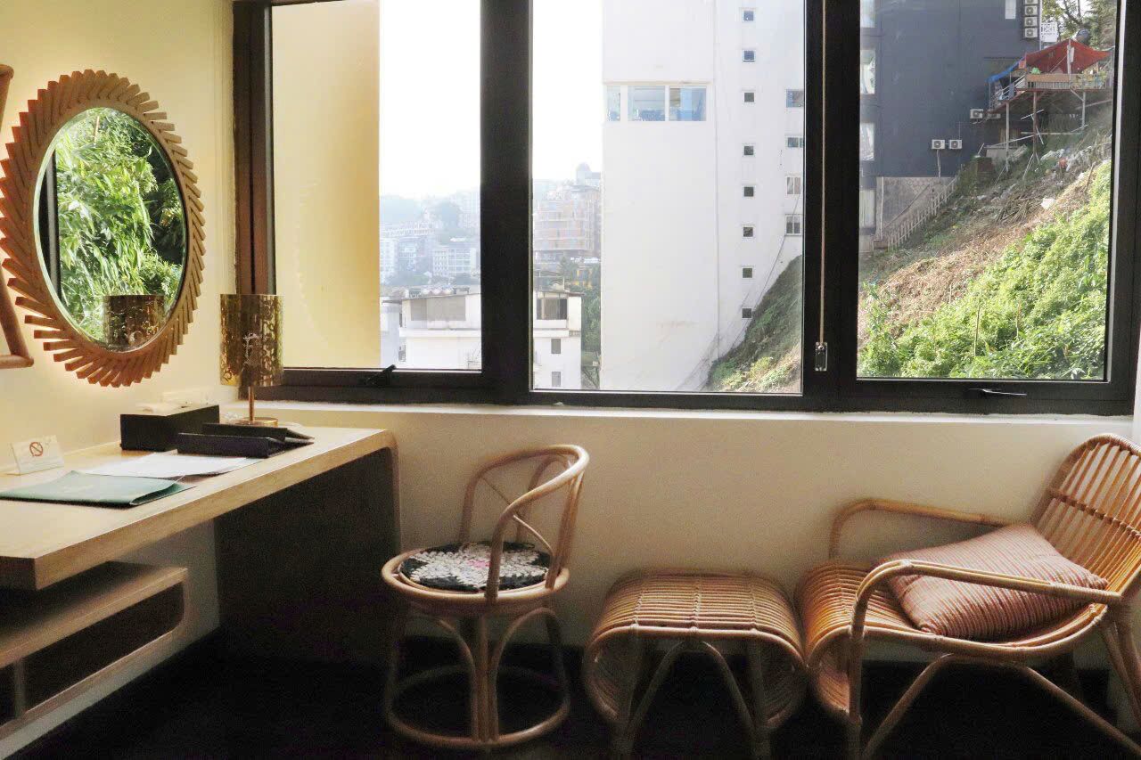 A room with a desk, two wicker chairs, a mirror, and a window with a view of buildings and greenery.