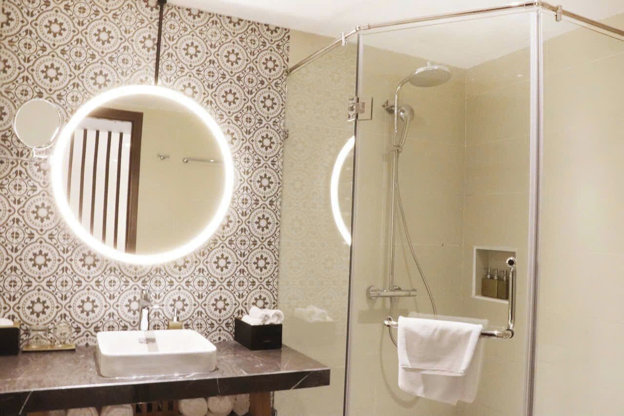 A bathroom with a glass-enclosed shower, a round mirror with a light, and a patterned wall.