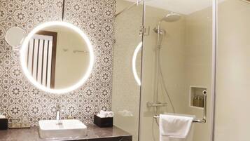 A bathroom with a glass-enclosed shower, a round mirror with a light, and a patterned wall.
