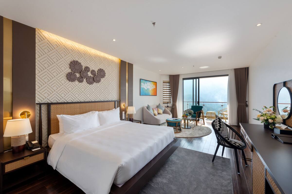 Habitación de hotel con una cama grande, un escritorio con silla, una mesita redonda y un balcón con vistas.