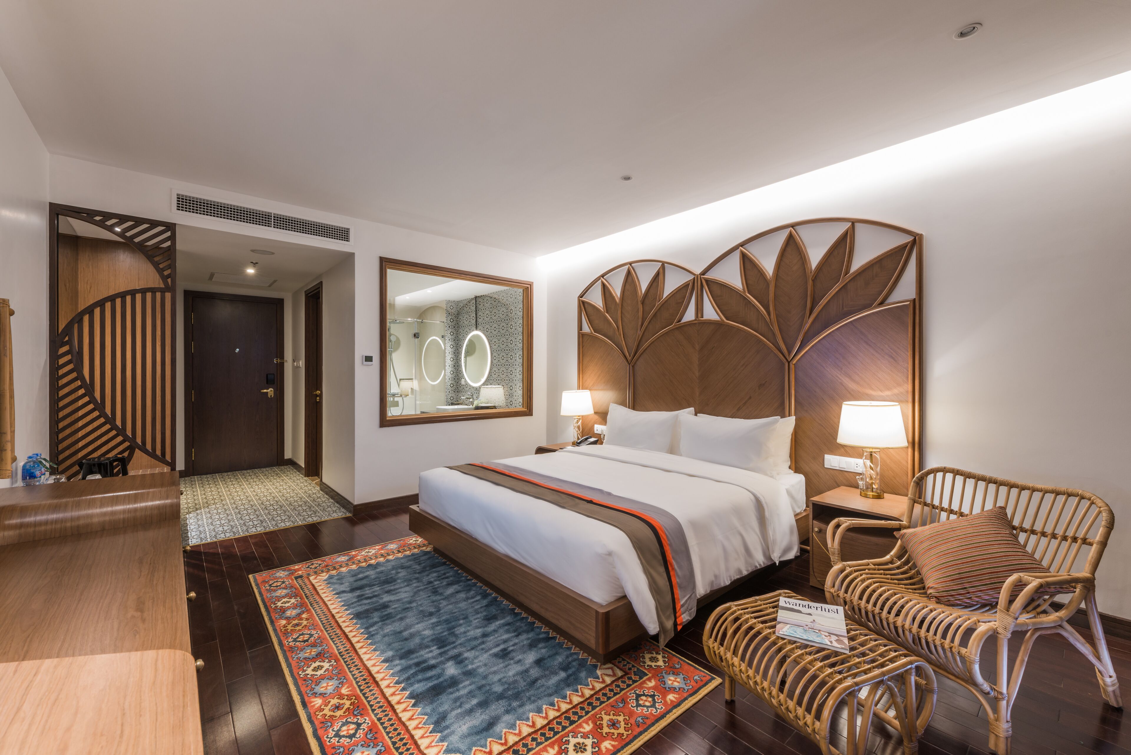 Une chambre d’hôtel avec un grand lit, une tête de lit en bois, une table de chevet, un fauteuil en osier et une petite table avec un magazine.