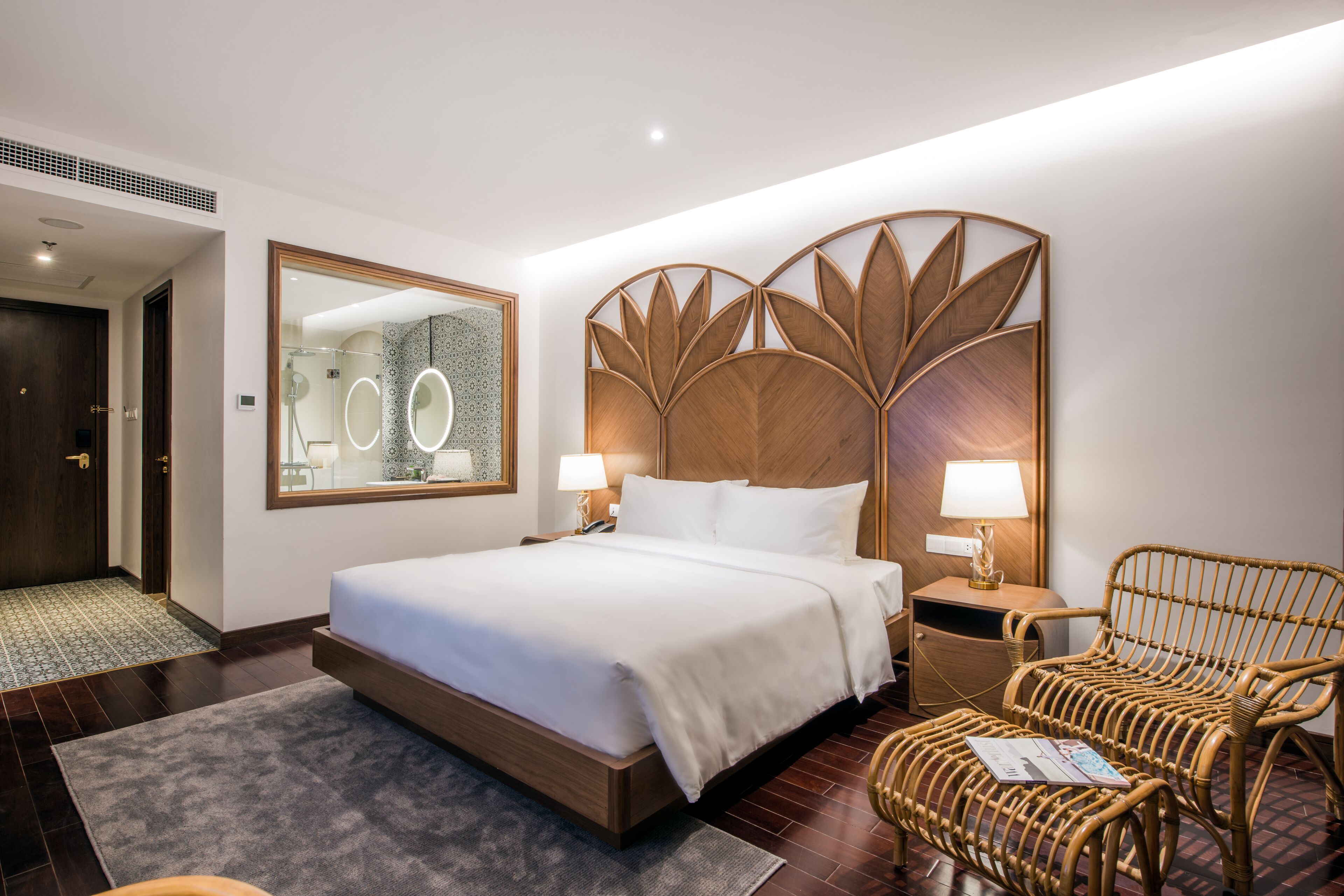 A hotel room with a large bed, a wooden headboard, a wicker chair, and a mirror with circular lights.