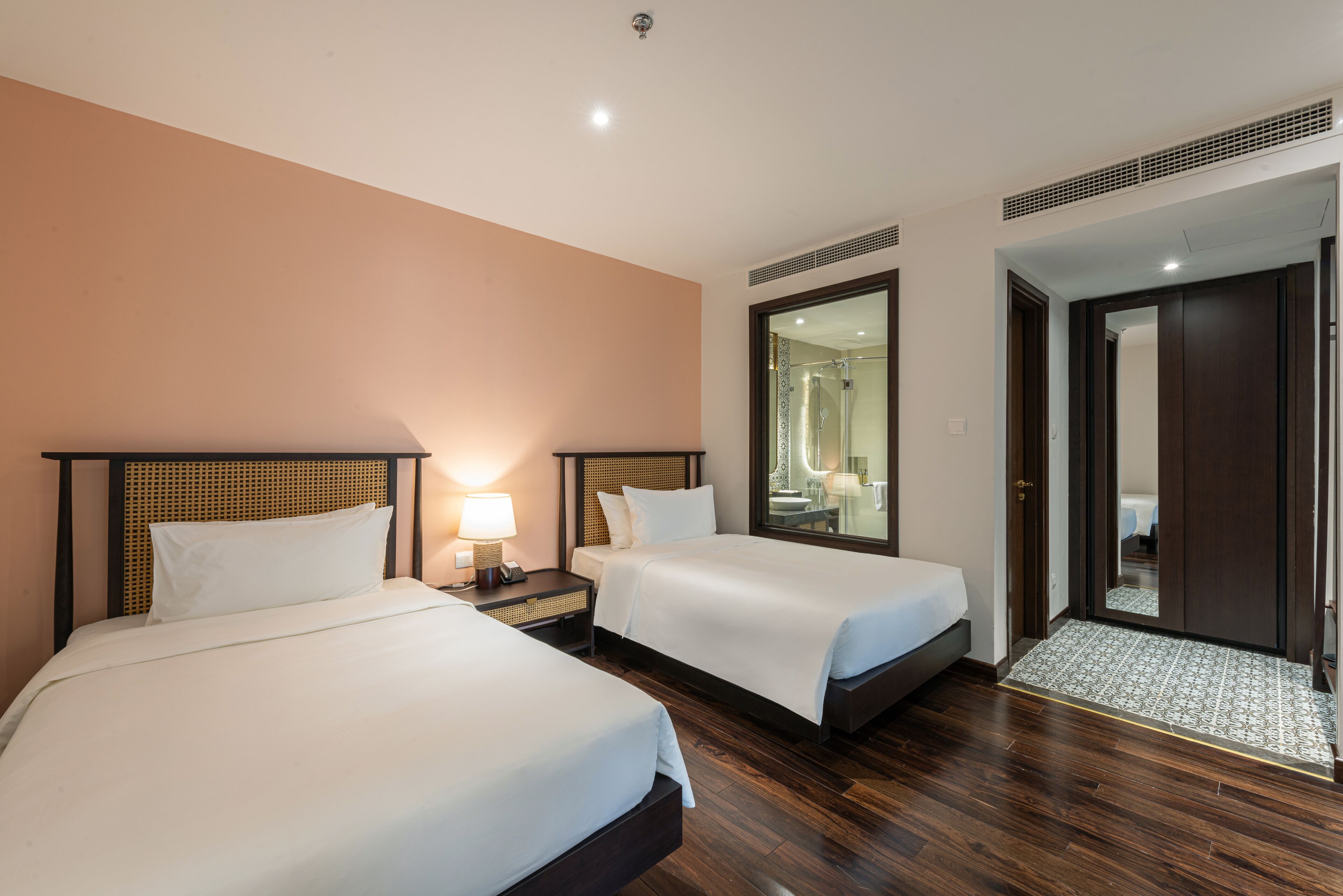Une chambre d’hôtel avec deux lits, une table de chevet avec une lampe, un grand miroir et un plancher en bois.