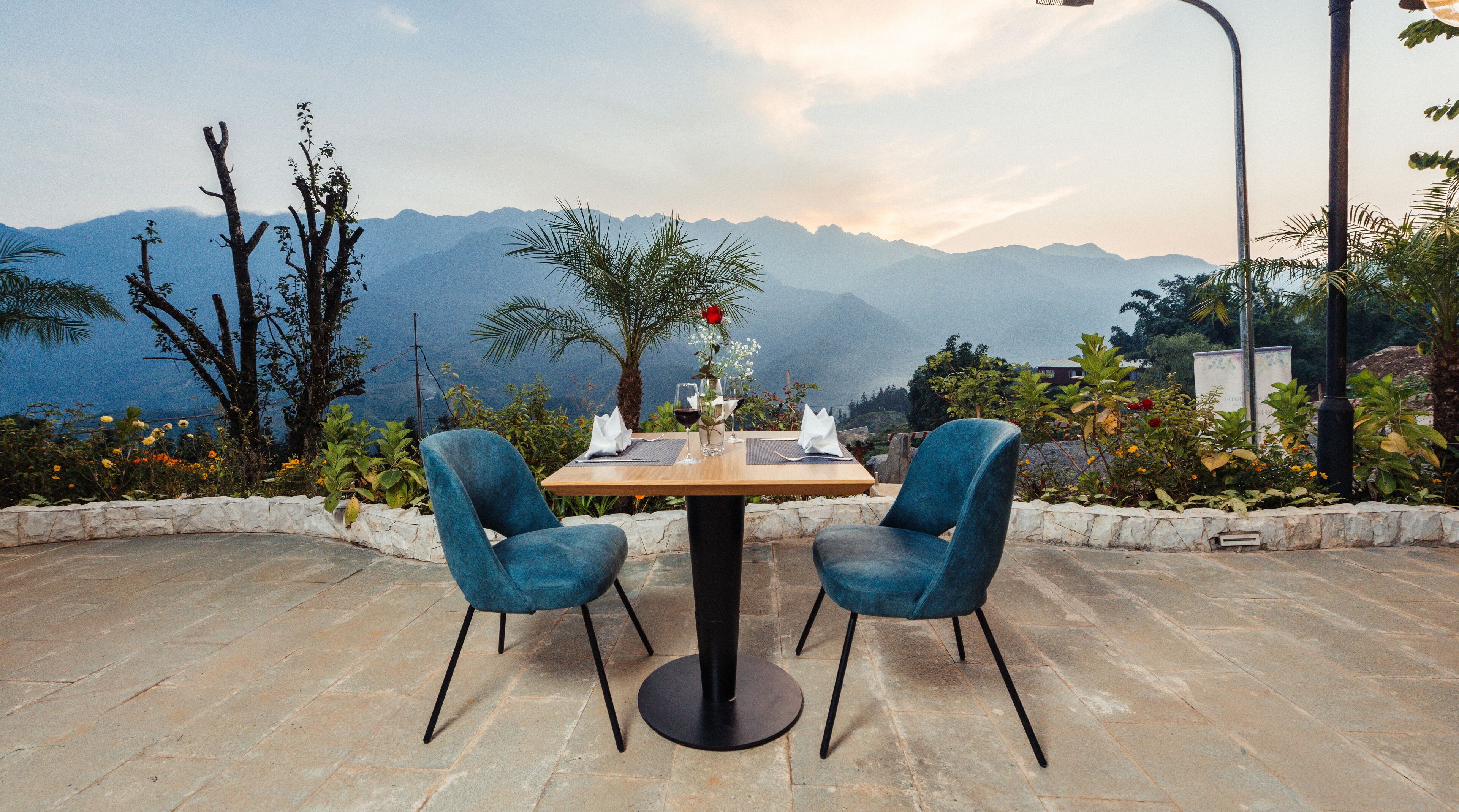 Una mesa para dos con vistas a la montaña, dos sillas azules y un florero con flores.