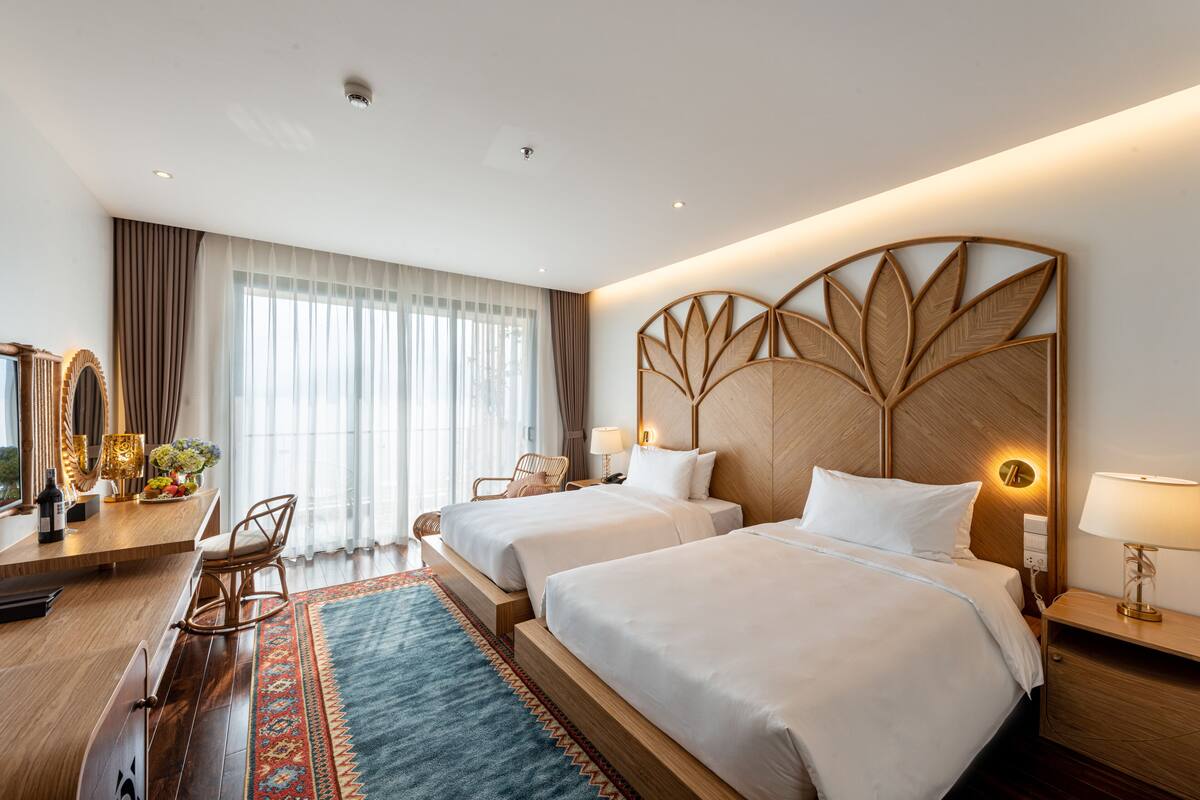 Habitación de hotel con dos camas, cabecero de madera, escritorio con silla, ventana grande con cortinas y un panel decorativo en la pared.