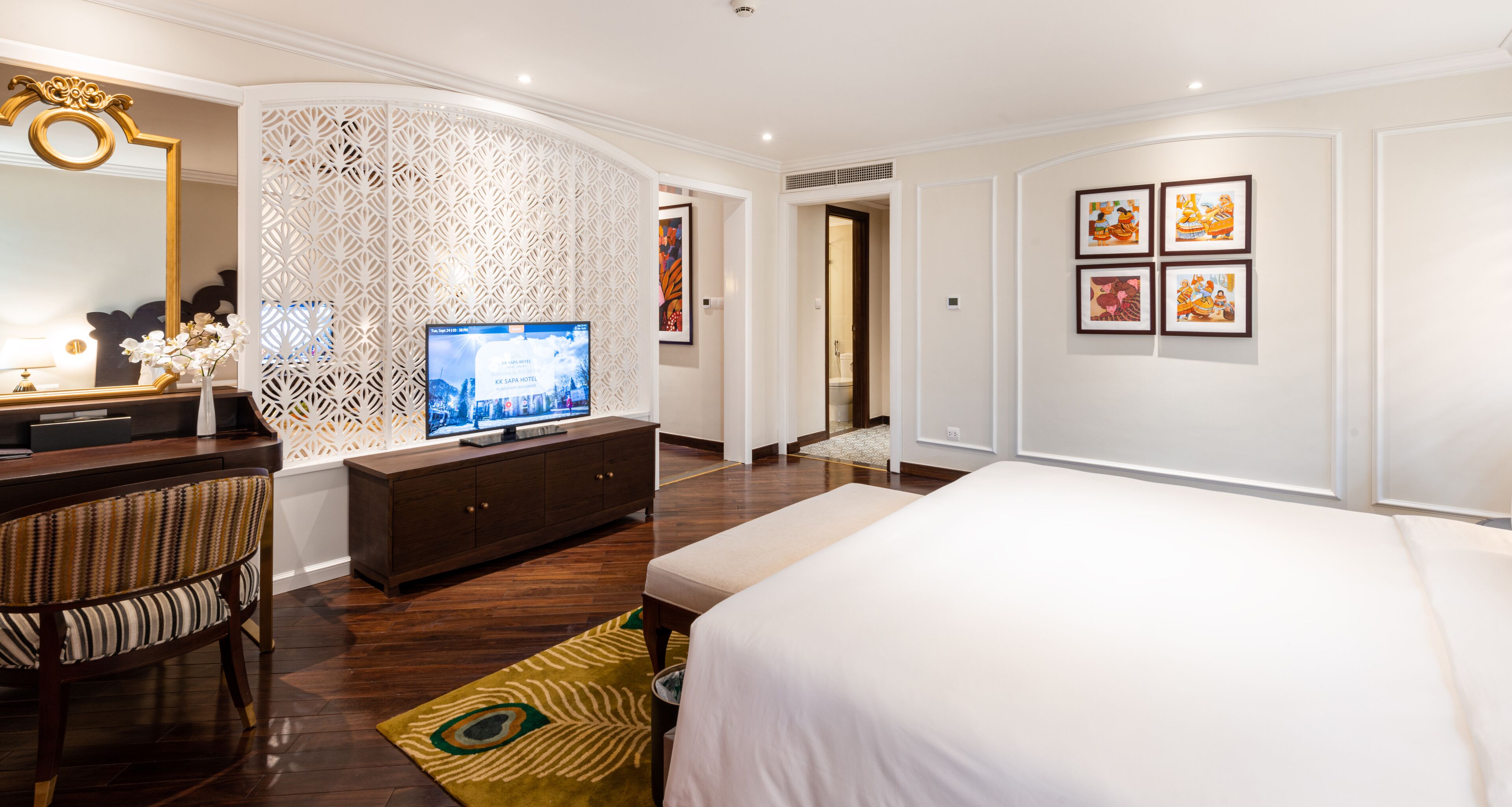 A modern hotel room with a large bed, a TV on a wooden console, a decorative wall panel, and framed artwork on the wall.