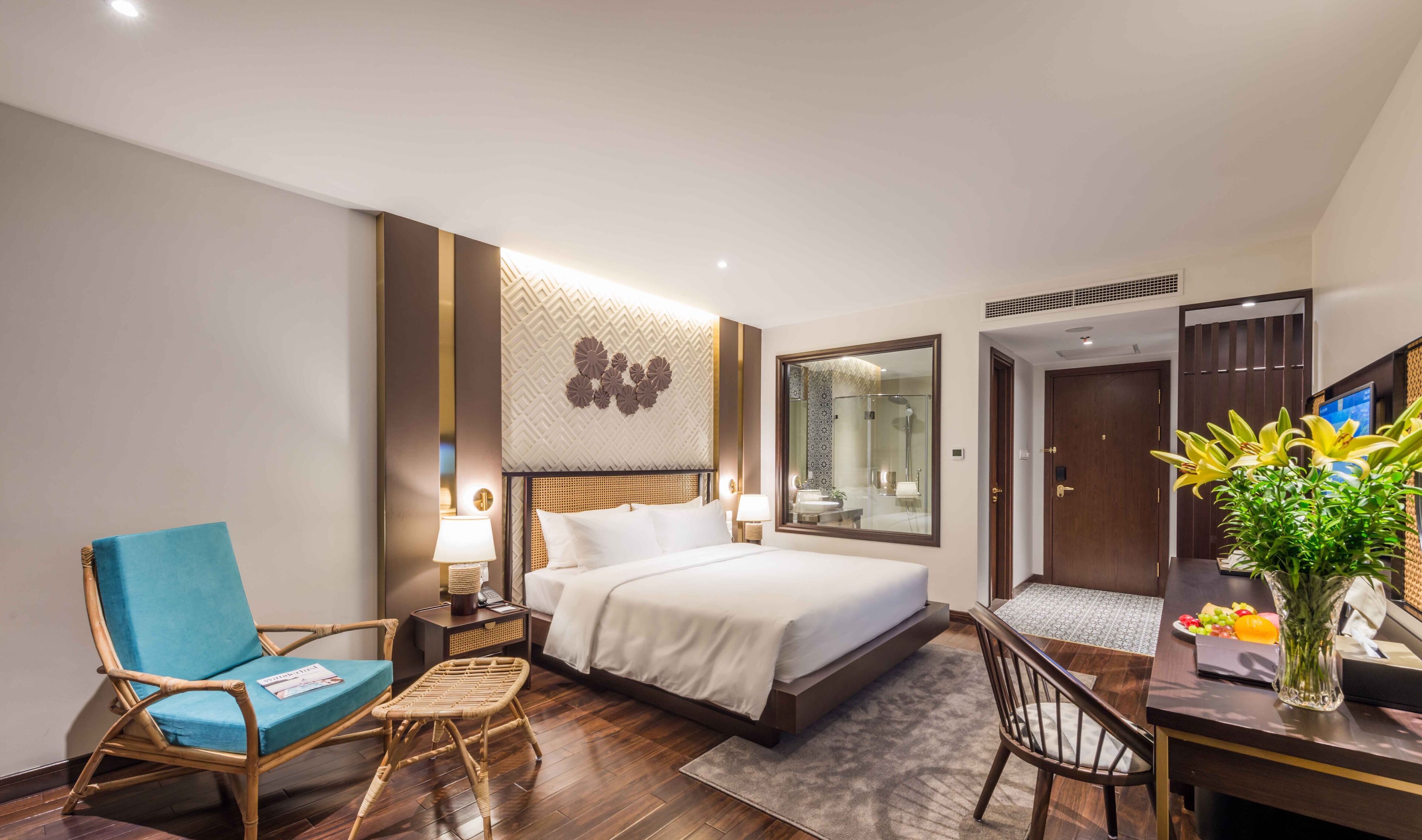 Une chambre d’hôtel comprenant un lit, un bureau, une chaise, une table de chevet, une lampe, un miroir, une porte et un vase avec des fleurs.