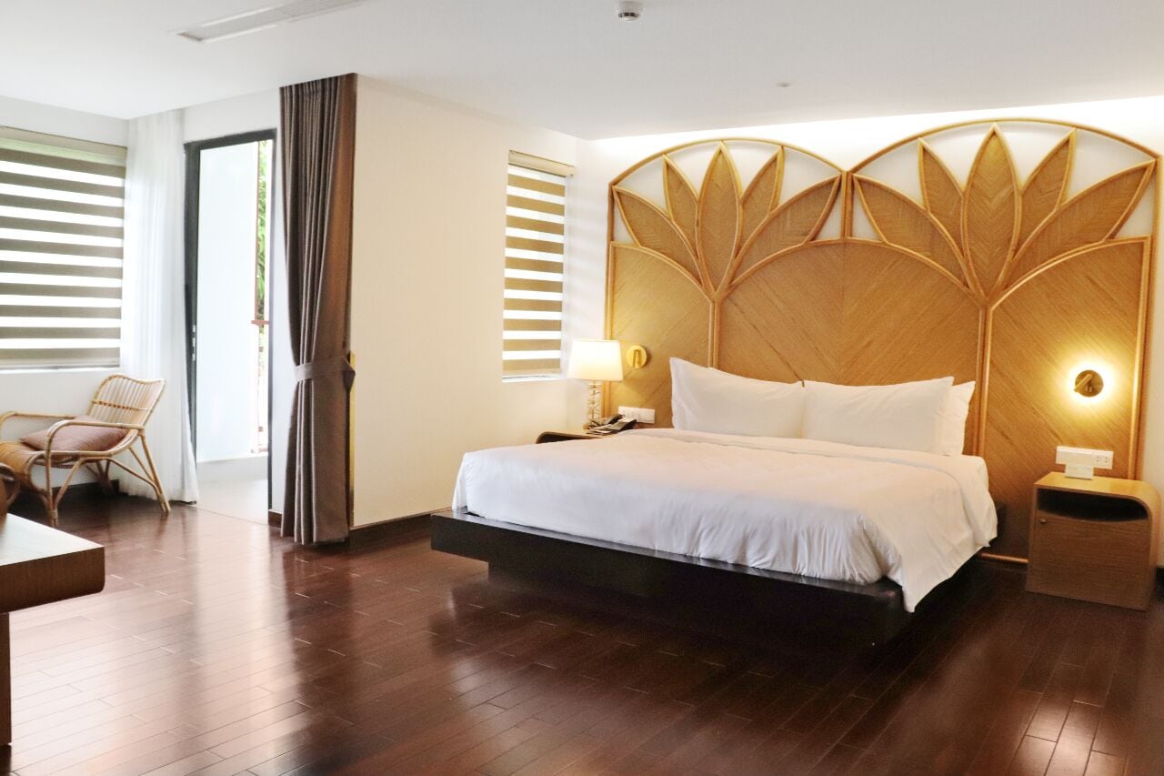 A modern hotel room with a large bed, a wooden headboard, a bedside table, a chair, and a view through a door.
