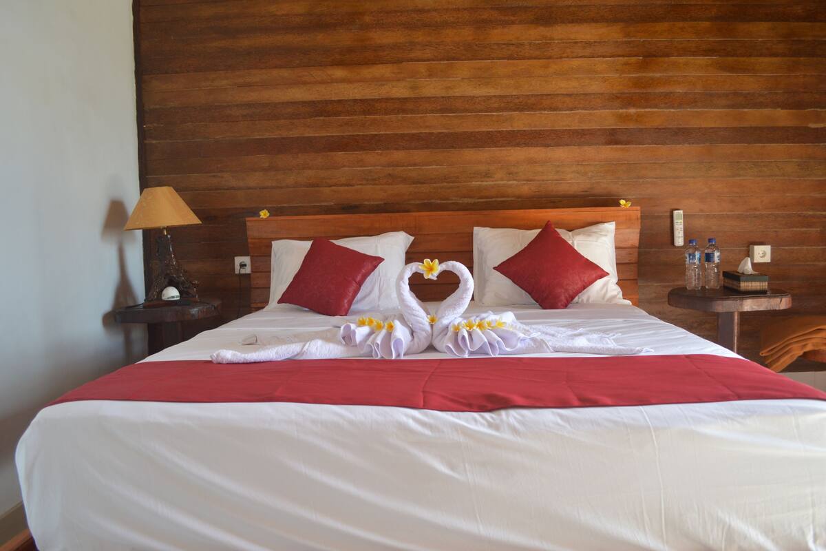 A hotel room with a wooden headboard, a bed with white linens, red pillows, and a red blanket. There are two nightstands with lamps and a table with water bottles.
