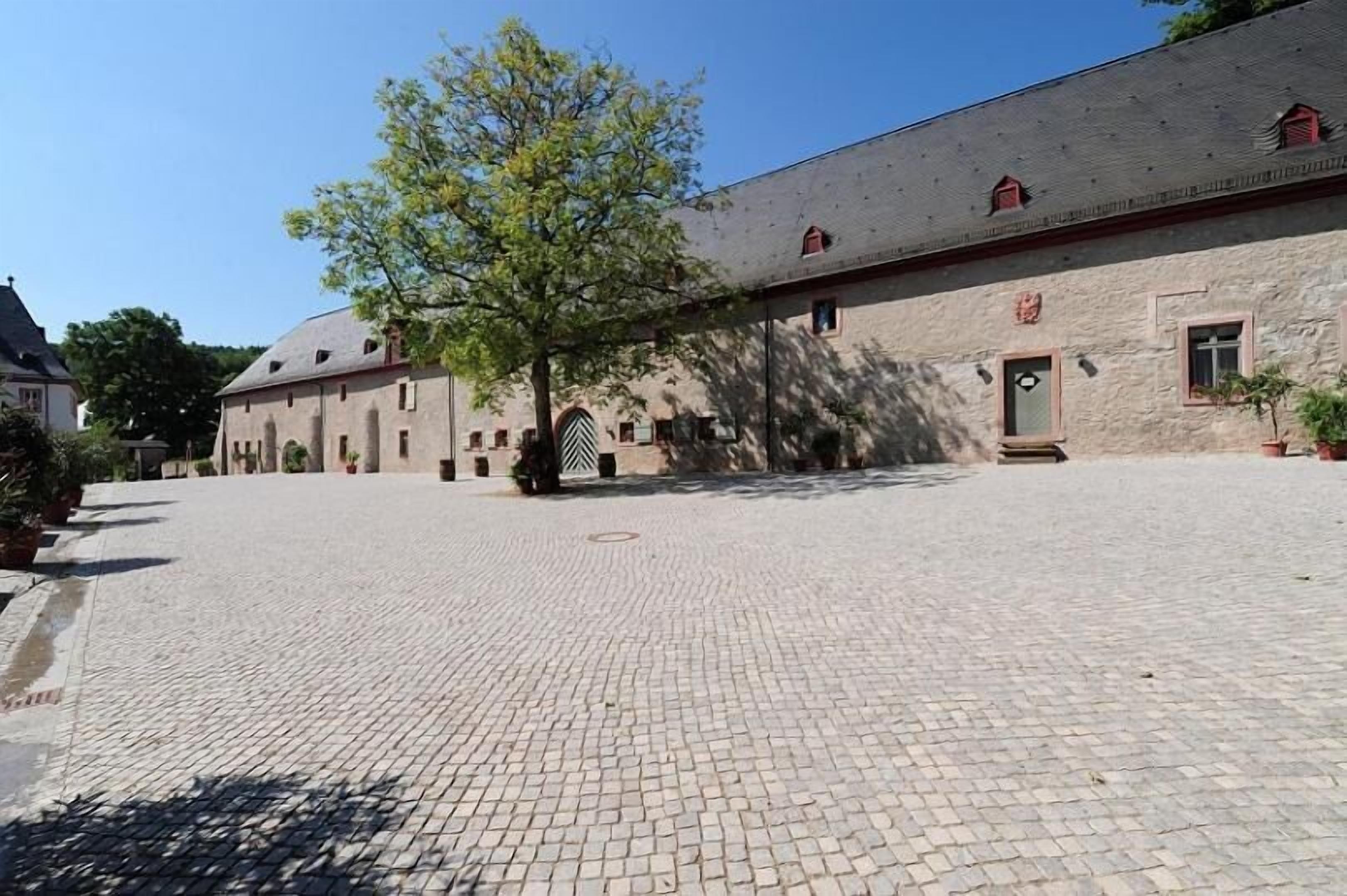 En innergård med kullerstensbeläggning, en stor byggnad med många fönster och ett träd som ger skugga.