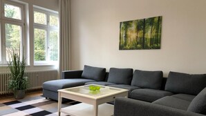 Una sala de estar moderna con un sofá seccional gris, una mesa de centro blanca y una alfombra a cuadros. Se ve una ventana con cortinas, un radiador y una planta en maceta.