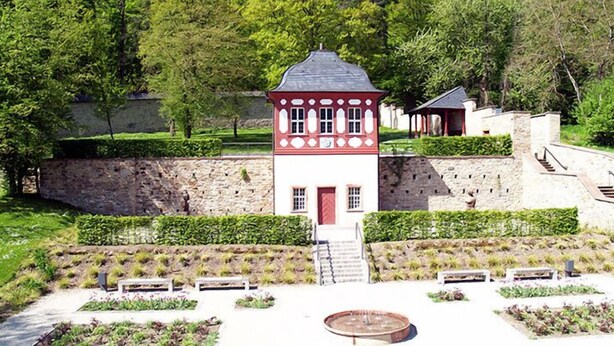 Una pequeña torre roja y blanca con una escalera que lleva a una puerta, rodeada de jardines cuidados y un muro de piedra.