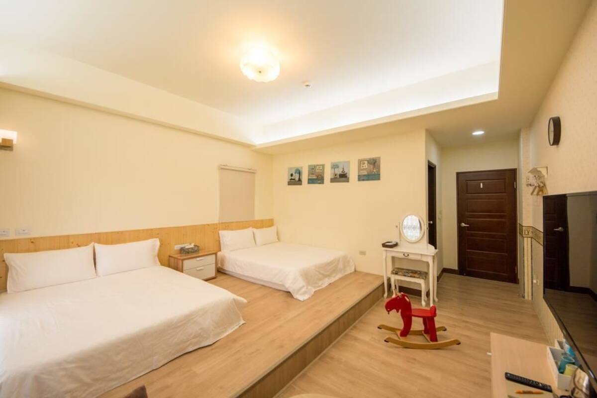 A hotel room with two beds, a wooden headboard, a small table with a mirror, a rocking horse, and a door leading to another room.
