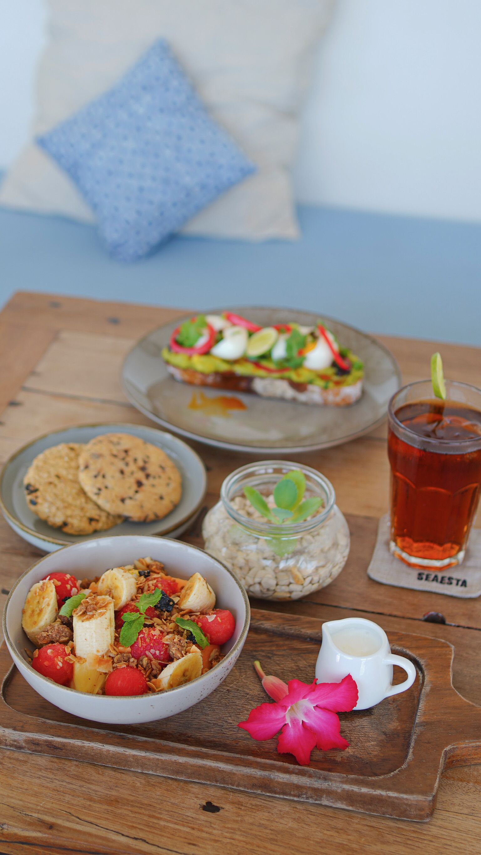 Un desayuno con ensalada de frutas, galletas, un vaso de té helado y una taza pequeña de leche sobre una bandeja de madera.