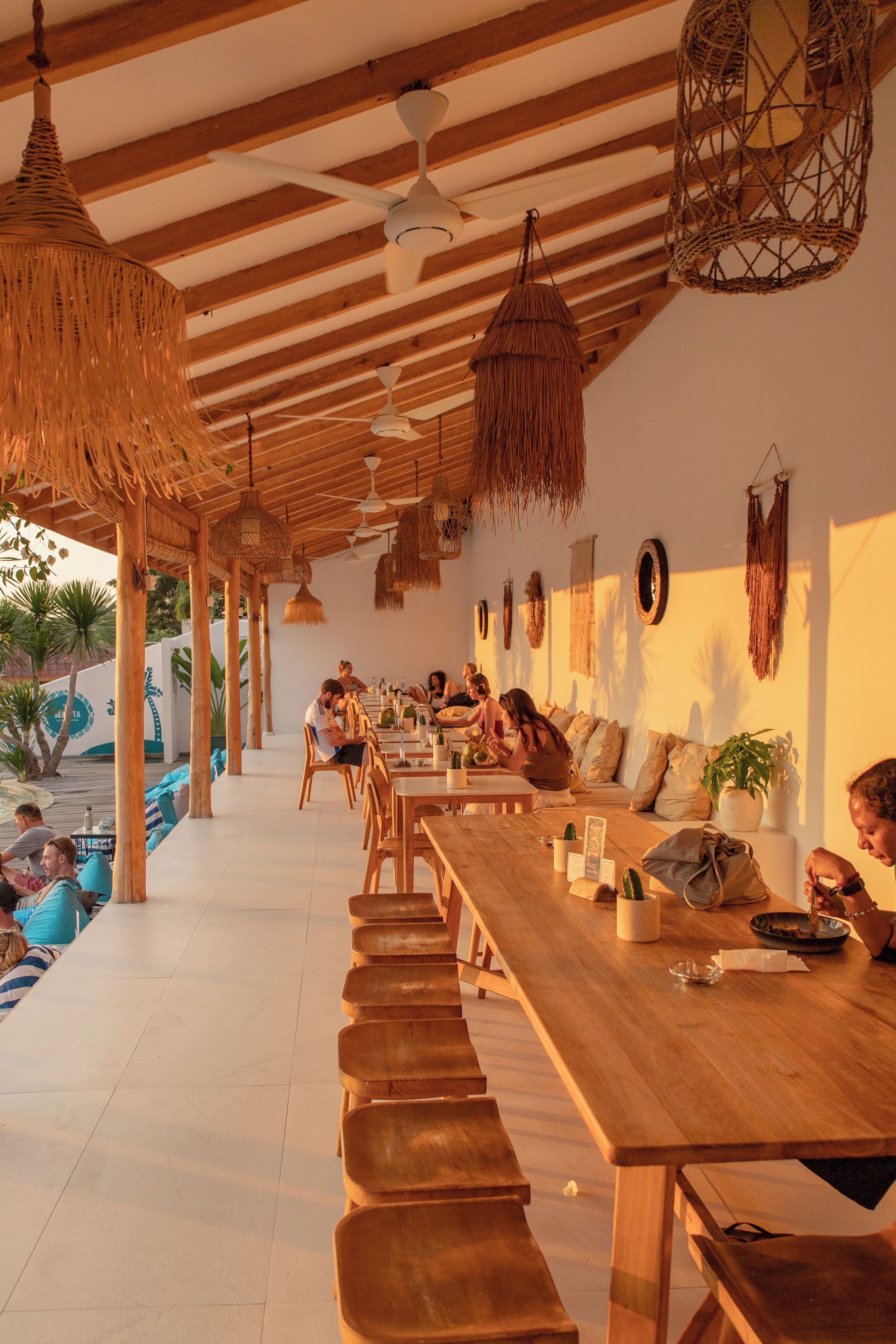 A restaurant with wooden tables and benches, hanging lights, and a pool area in the background.
