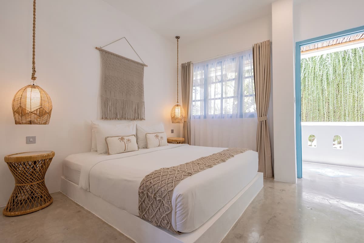 Ein geräumiges Schlafzimmer mit einem großen Bett, zwei Nachttischen, einer Wanddekoration aus Makramee und Blick auf Grün durch das Fenster.