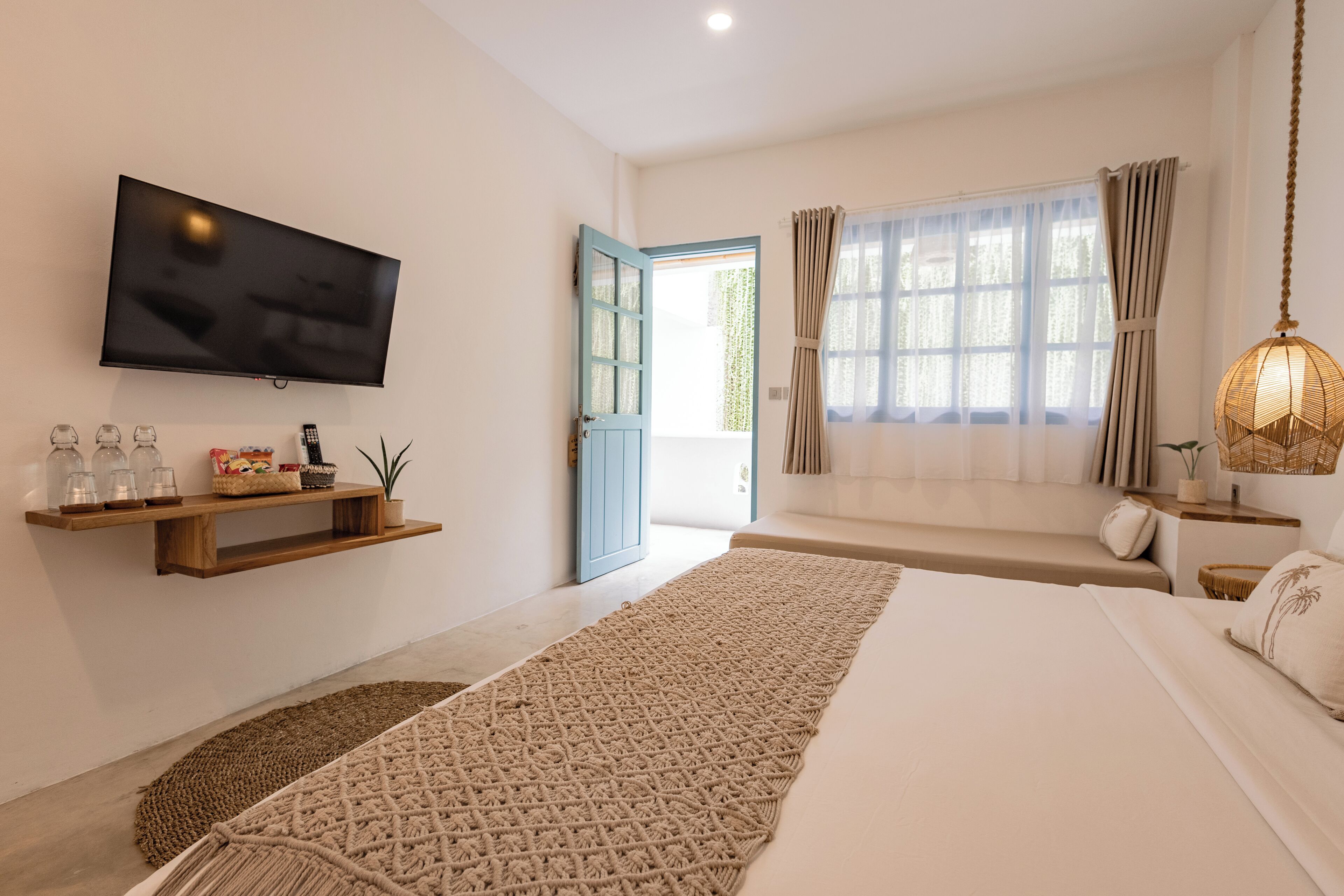 Une chambre moderne avec un grand lit, un téléviseur fixé au mur, une étagère en bois ornée d’objets décoratifs, une grande fenêtre avec des rideaux et un luminaire suspendu.