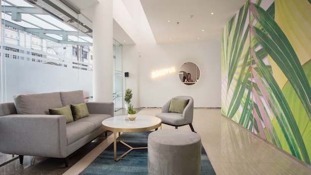 Un hall d’entrée moderne avec un canapé gris, une table basse ronde et un pouf gris. On peut y voir un grand miroir et un mur orné d’un motif abstrait vert et jaune.