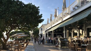 An outdoor cafe with tables and chairs, a tree, and a building with a balcony.