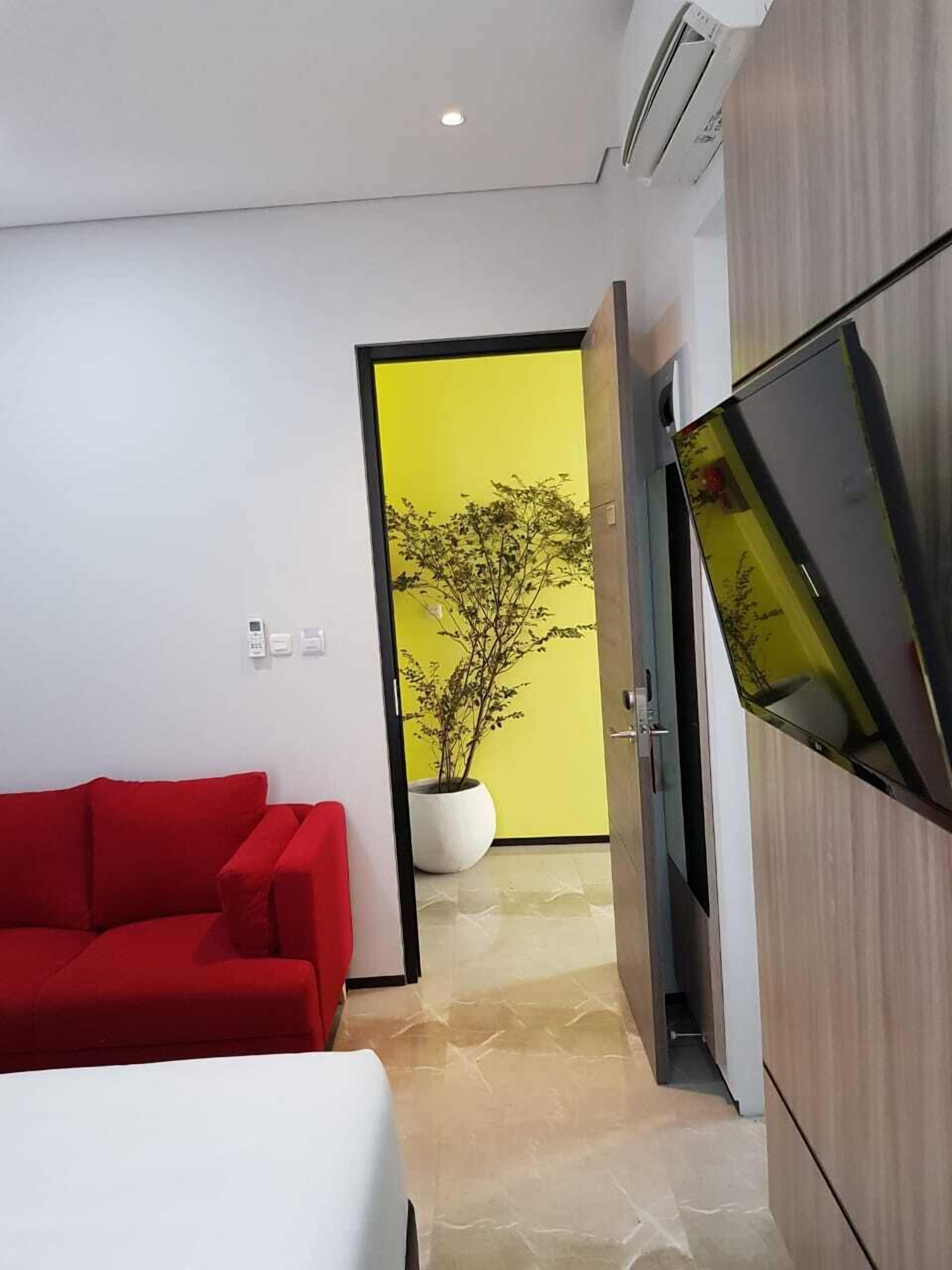 A modern hotel room with a red sofa, a bed, a flat-screen TV, and a potted plant in the corner. - POD House Pakubuwono - Hostel (Jakarta)
