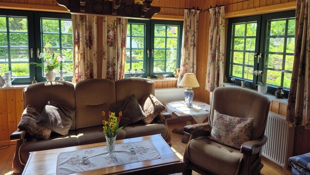 A cozy room with wooden walls, a chandelier, a table with a vase of flowers, and large windows with curtains. - Cozy wooden cottage with Wi-Fi and feel-good atmosphere (Bohmte)