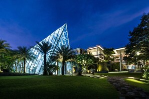 Un edificio moderno con una estructura piramidal de vidrio, rodeado de follaje cuidado y un camino de piedra.
