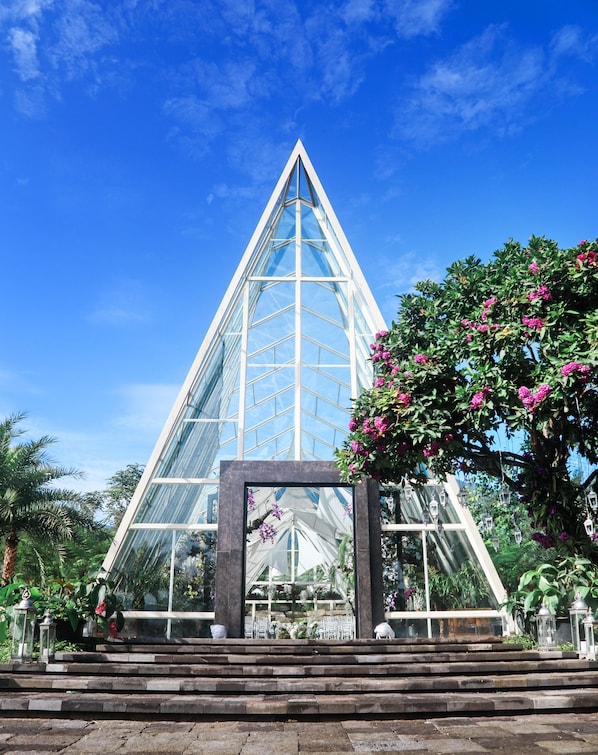 Una gran estructura piramidal de vidrio con un interior reflectante, rodeada de abundante vegetación y flores rosadas.