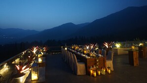 An outdoor dining area with a long table set for a meal, surrounded by chairs, and decorated with candles and plants.