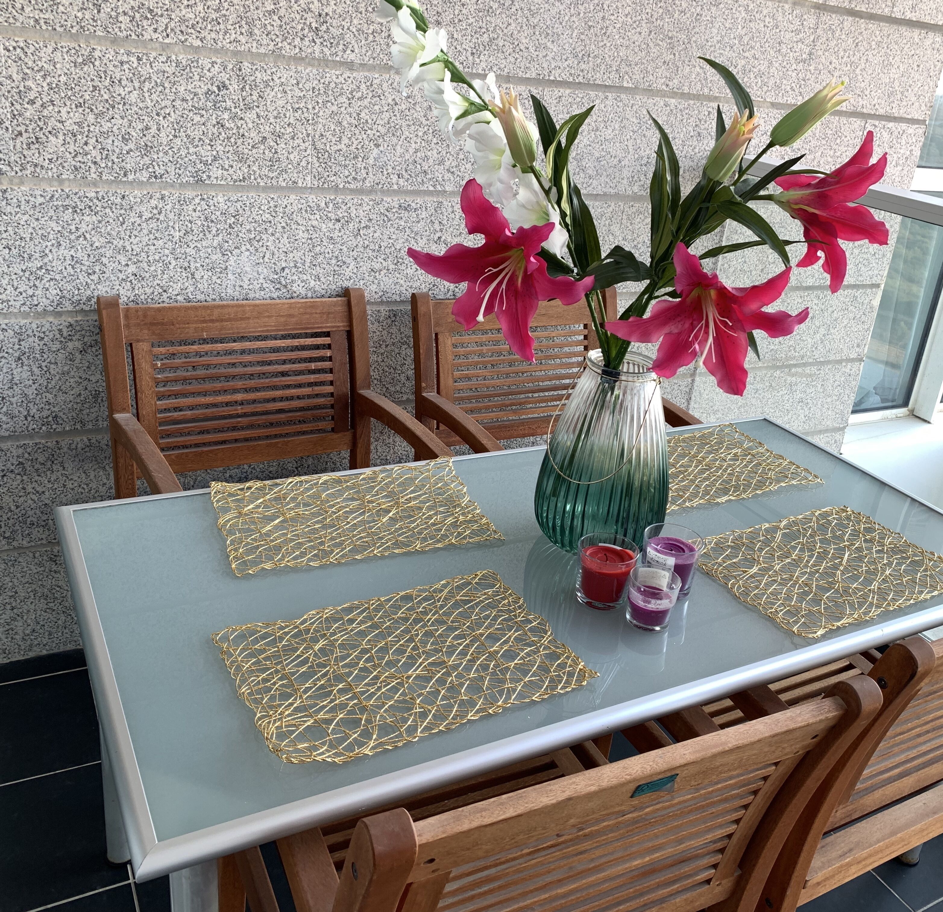 A table with a clear glass top, surrounded by wooden chairs. A vase with pink and white flowers is placed on the table.
