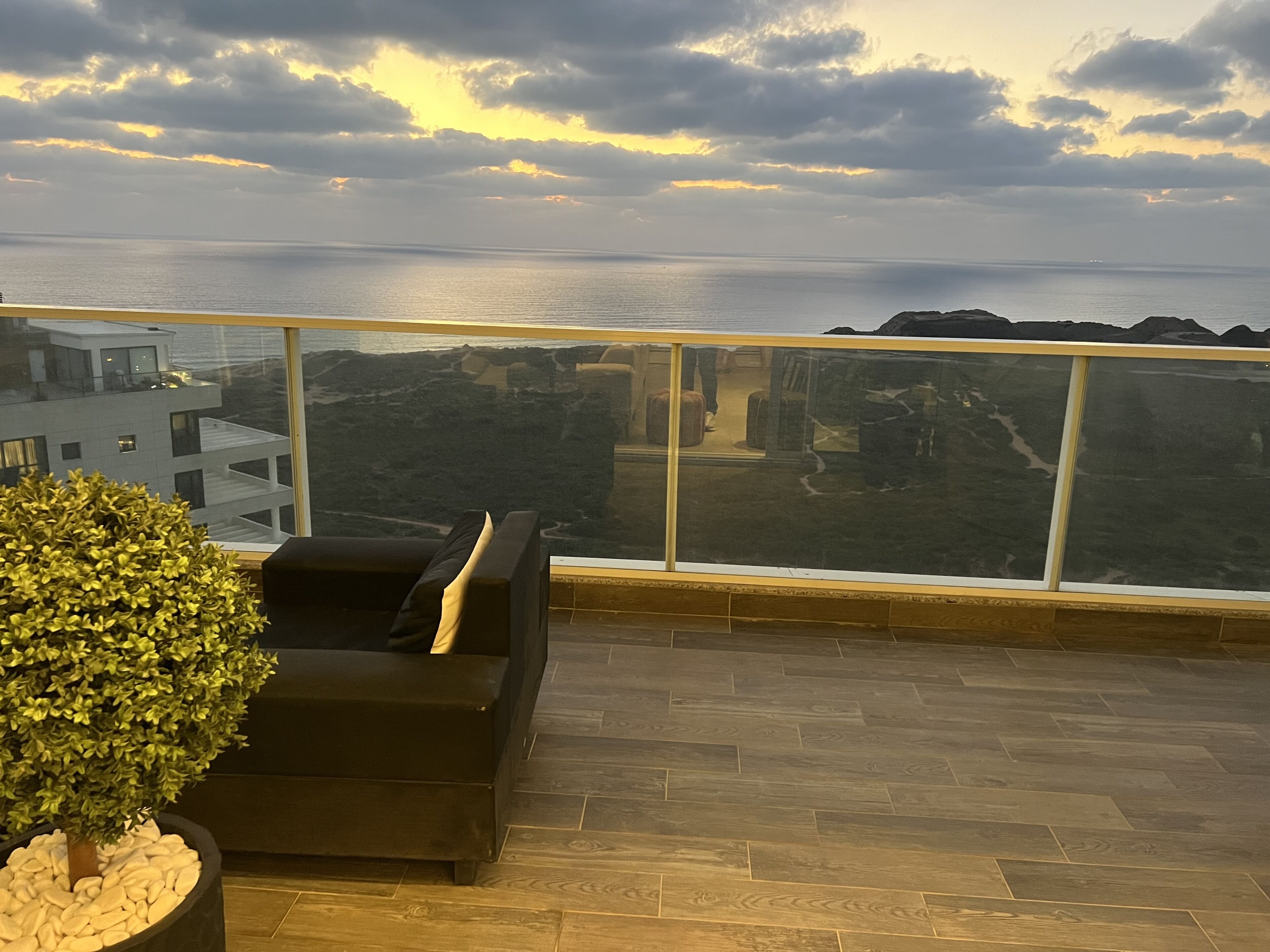 Balcón con sofá negro y planta en maceta, con vistas a un paisaje costero con edificios y una masa de agua.