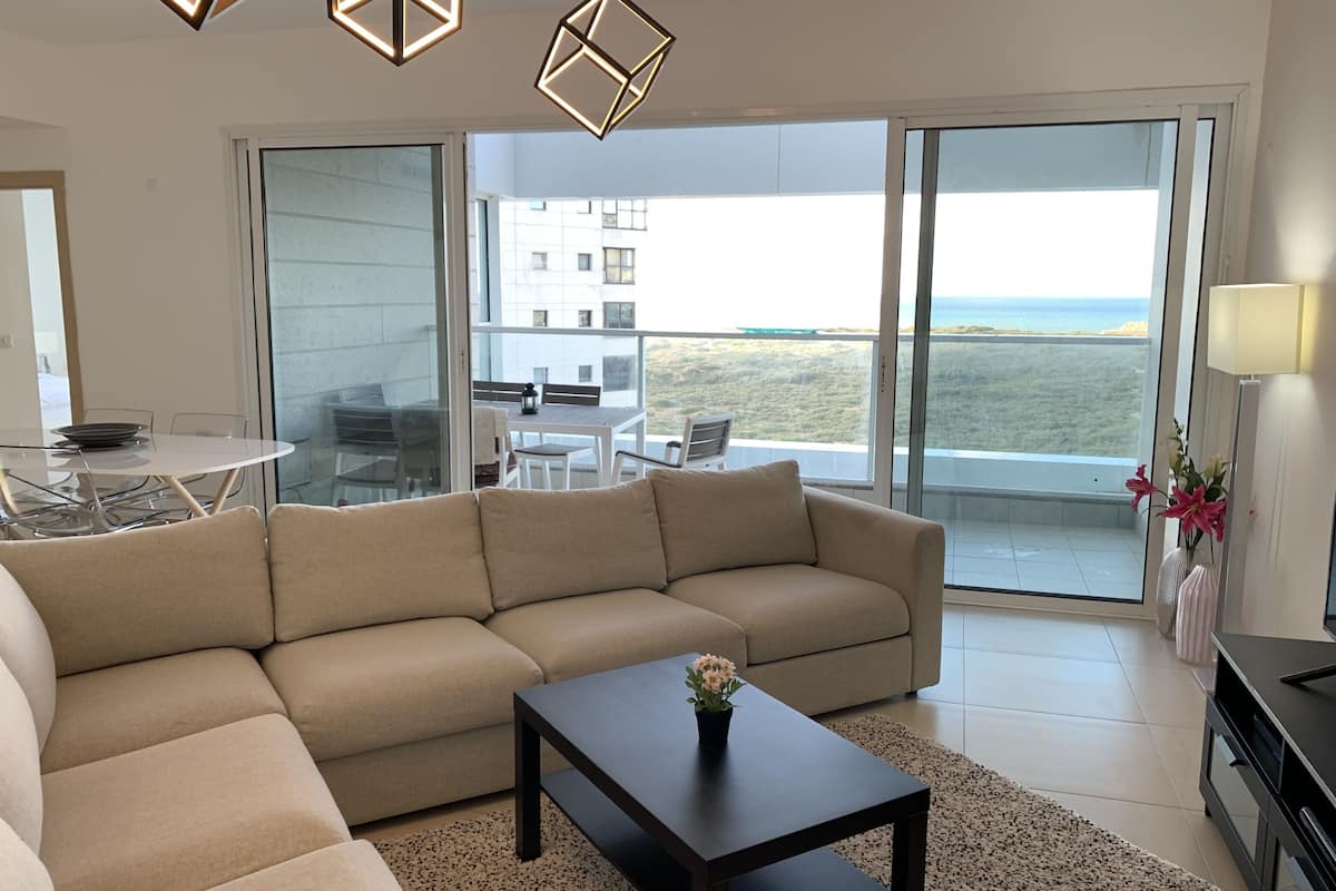 Ein modernes Wohnzimmer mit einem beigen Ecksofa, einem dunklen Couchtisch und einem Glastisch. Ein Balkon bietet Blick auf das Meer und Gebäude.