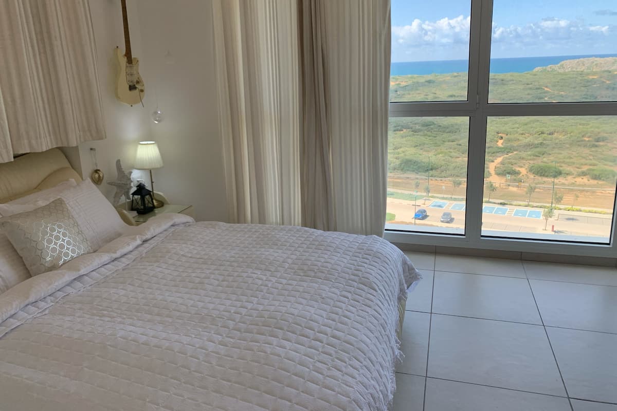 Ein Schlafzimmer mit Bett, Vorhängen, einer Gitarre, einem Nachttisch und Blick auf eine Landschaft durch ein großes Fenster.