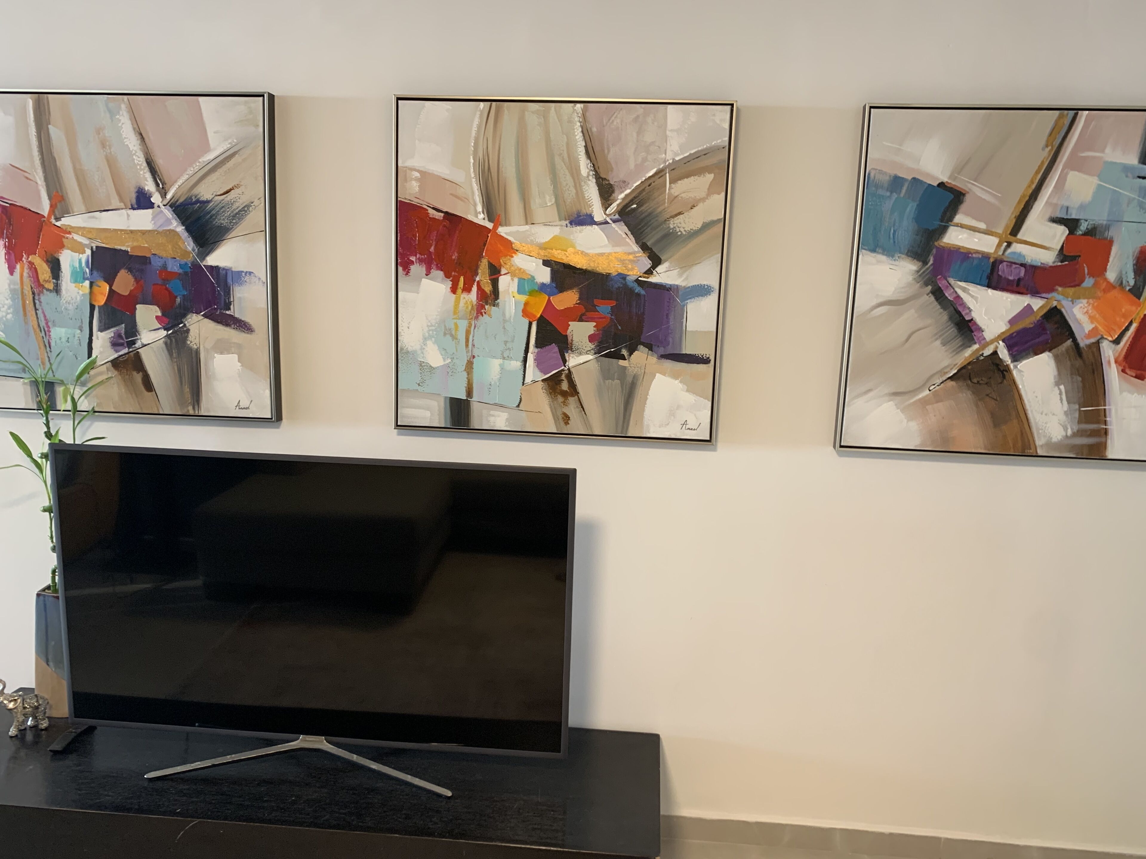 A wall with three abstract paintings and a flat-screen TV on a stand.