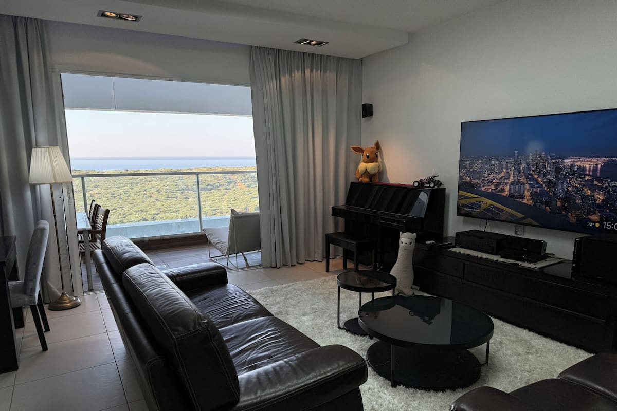 Ein modernes Wohnzimmer mit einem großen Fenster, einem schwarzen Sofa, einem Klavier und einem Fernseher, der eine Stadtansicht zeigt.