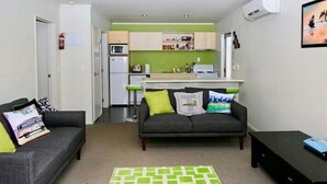 Un espacio habitable compacto con un sofá gris, un tapete verde con estampado y una pequeña mesa negra. Se aprecia una kitchenette con armarios blancos y un microondas al fondo.