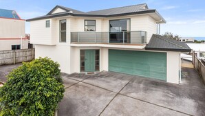 Une maison à deux étages avec une porte de garage verte, un balcon et un petit jardin devant la maison, agrémenté d’un arbuste.
