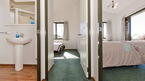 A hotel room with a large bed, a bedside table, a window with curtains, a bathroom with a sink and mirror, and a hallway leading to another room.