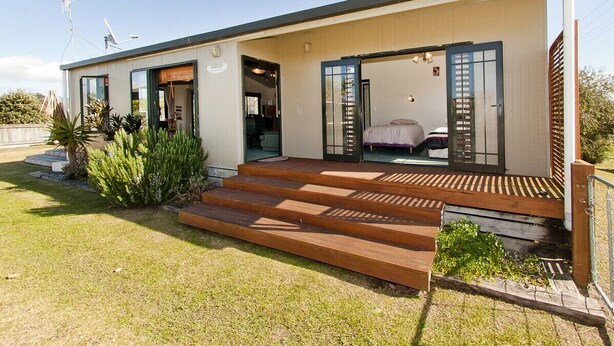 A modern house with a flat roof, a front porch with wooden steps, and a visible bedroom with a bed.