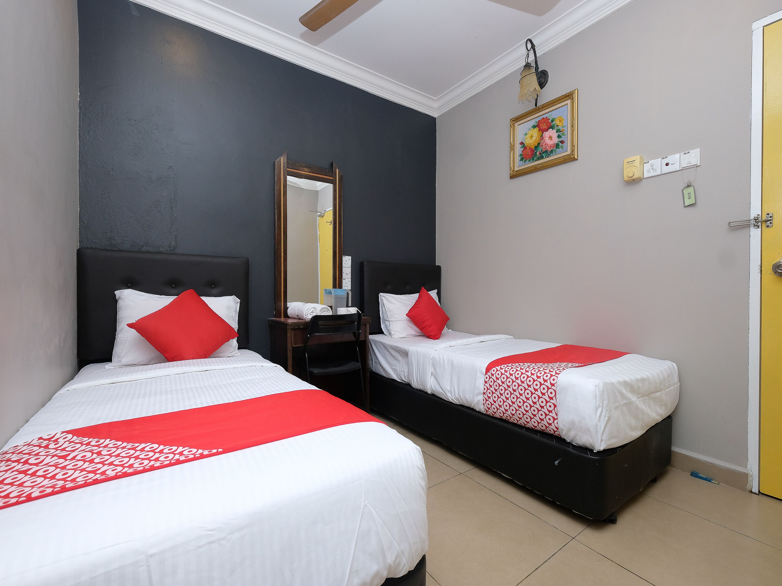 A hotel room with two beds, each with white bedding and red accents, a wooden nightstand, a mirror, and a painting on the wall.