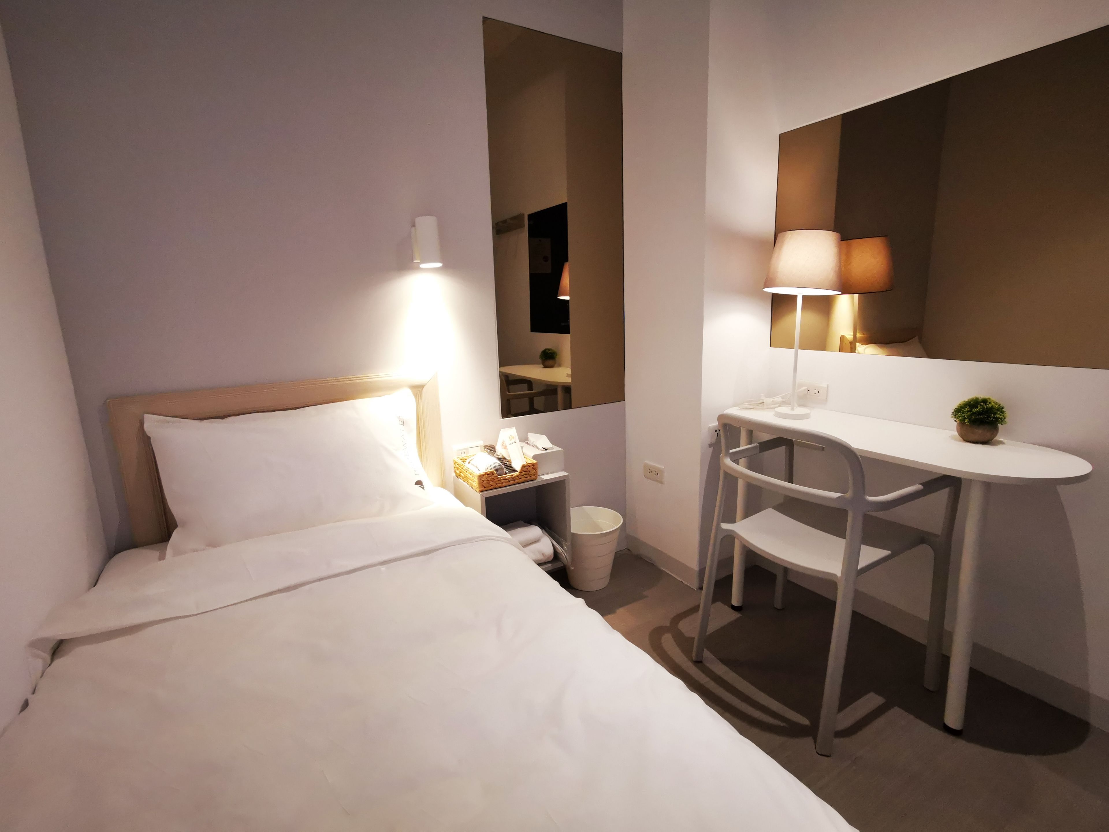 Habitación de hotel con cama, mesita de noche, lámpara, espejo y un escritorio pequeño con silla.