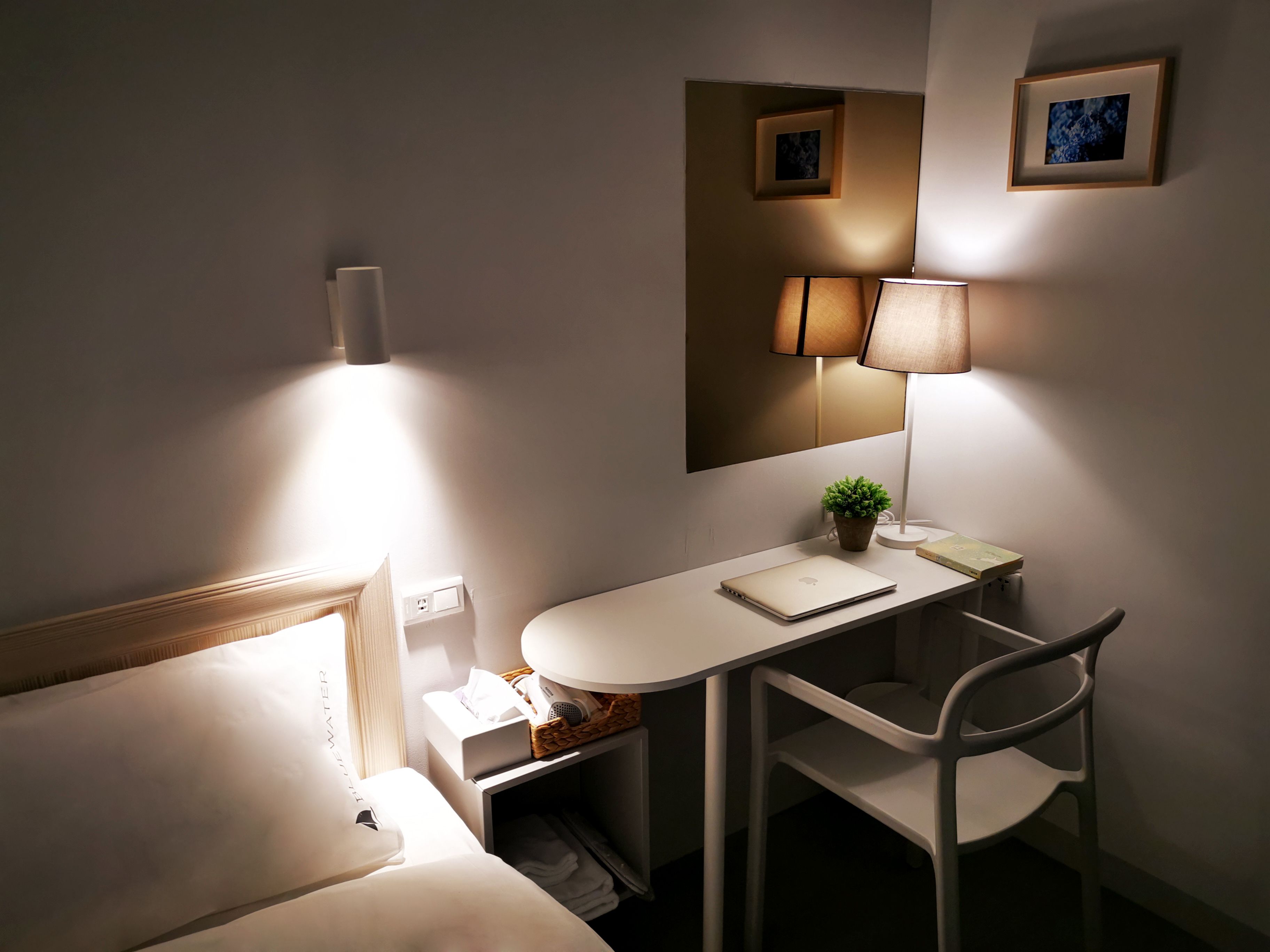 A hotel room with a bed, a desk with a laptop, a chair, a lamp, a small plant, and a mirror.