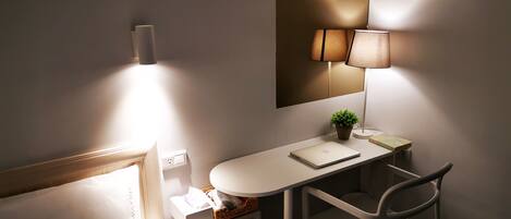 A hotel room with a bed, a desk with a laptop, a chair, a lamp, a small plant, and a mirror.