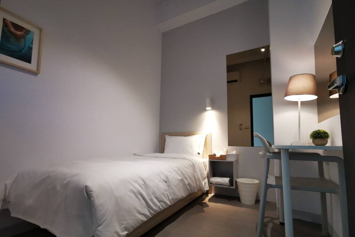Un dormitorio con cama, mesita de noche, lámpara, silla y una pequeña planta en maceta.