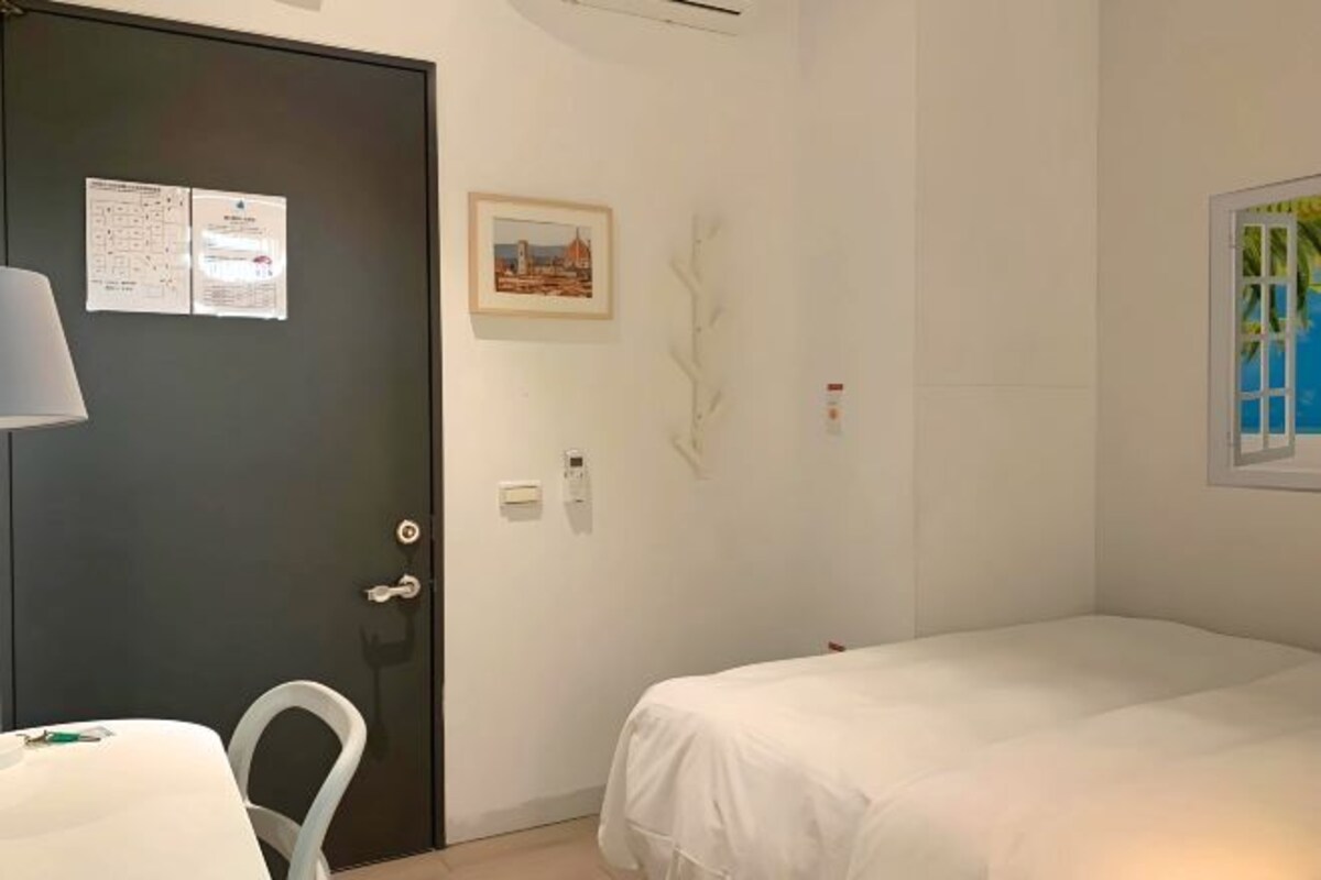 Hotellihuone, jossa on sänky, työpöytä tuolilla, ovi, jossa on ilmoitustaulu, seinään asennettu valokytkin ja kehystetty kuva.