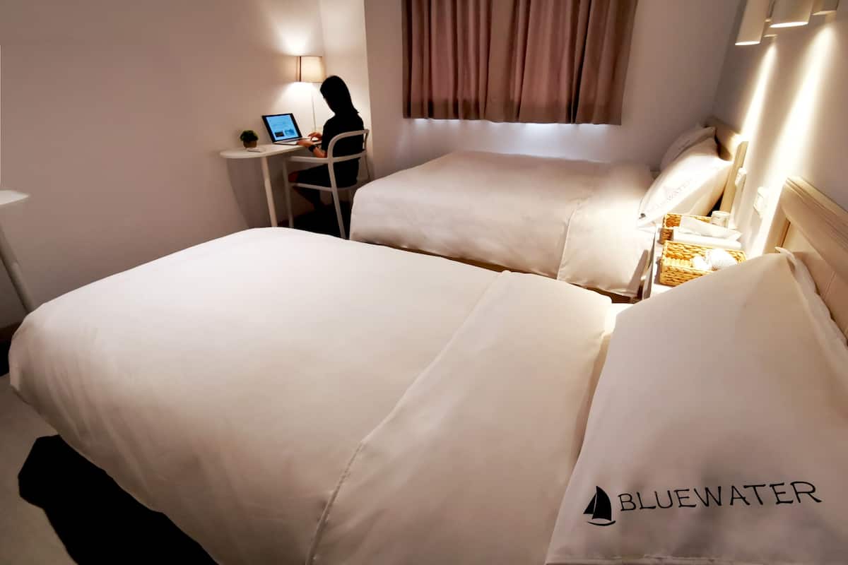 Una habitación de hotel con dos camas, un escritorio con una computadora portátil, una lámpara y una ventana con cortinas.